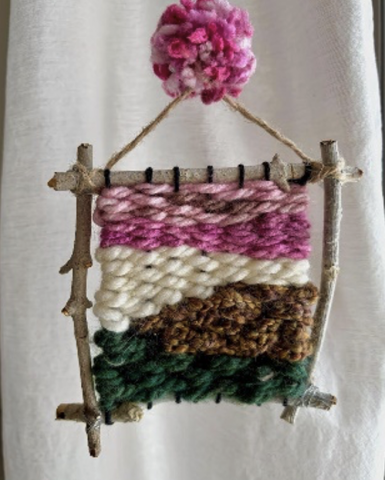 A frame of 4 sticks woven together with green, brown, white, and pink fibers.