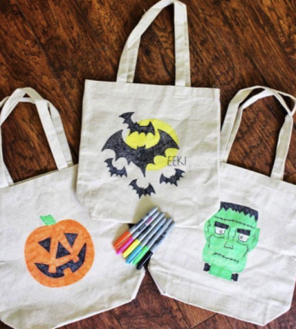 3 tote bags laying on a wooden floor, the first with an orange jack-o-lantern drawn on it, the middle with a yellow moon and 5 bats flying in front of it with the words EEK! written on it, and the last one with a green Frankenstein's monster drawn on it. In the middle of the 3 totes is an orange, pink, yellow, blue, green, and black sharpie. 