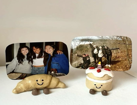A clay croissant with a smiley face and a clay strawberry cake with a smiley face holding two pictures of a group of girls.