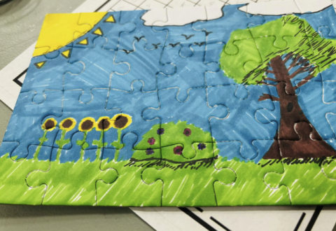 Puzzle with grass, a shrub, a tree, a sun, two clouds, and five sunflowers drawn on it with markers.
