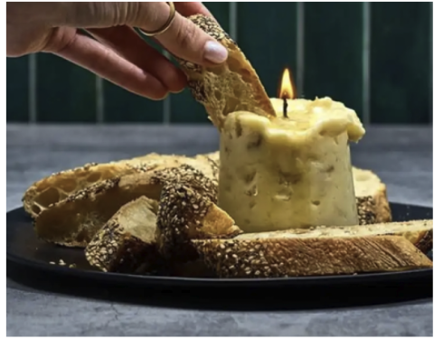 Image of a lit candle made out of butter with a person's hand showing bread being dipped into the melting butter.