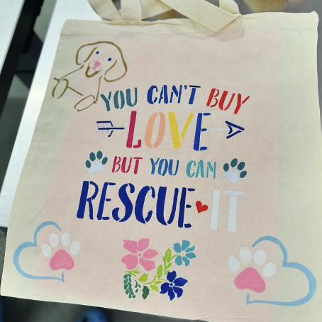 A tote bag with a puppy, paw prints, flowers, and the words "YOU CAN'T BUY LOVE BUT YOU CAN RESCUE IT" stenciled in paint on the bag.