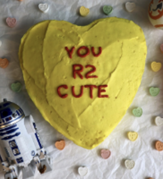 A yellow conversation heart shaped cake with the words "YOU R2 CUTE" iced on it in red. There is a mini R2-D2 from Start Wars in the bottom left corner of the image as well.