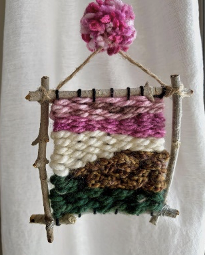 A frame of 4 sticks woven together with green, brown, white, and pink fibers.