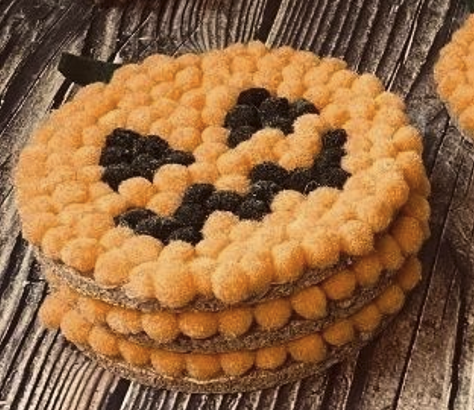 Pumpkin coaster made out of orange and black pom poms on a wooden table, stacked 3 pumpkins high.